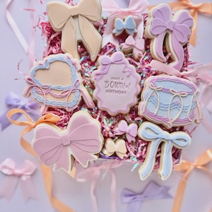 Könnte beinhalten: Eine Schachtel mit schleifenförmigen Keksen in Pastellfarben, darunter Rosa, Pfirsich und Blau, mit dekorativem Zuckerguss. Einige Kekse tragen den Text "Have a Bowtiful Birthday". Bänder in verschiedenen Farben umgeben die Schachtel.