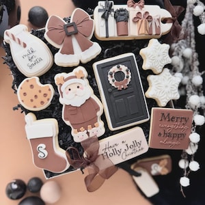 Könnte beinhalten: Eine Sammlung dekorierter Weihnachtsplätzchen in einer Geschenkbox. Die Kekse zeigen eine Milchflasche, eine Schleife, Geschenkboxen, Schneeflocken, eine Tür mit einem Kranz, einen Weihnachtsmann, einen Strumpf und einen Keks mit dem Text "Merry everything & happy always".