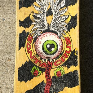 May include: A vintage skateboard deck with a yellow and black striped design. The deck features a graphic of a red eyeball with a green iris, a red tongue, and the words "SANTA CRUZ SKATEBOARDS" in a circular design.