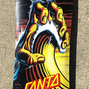 May include: A skateboard deck featuring a graphic of a large hand reaching over a wave with a surfer. The hand is yellow and orange, the wave is yellow and white, and the background is black with red accents. The words "SANTA CRUZ" are in red and yellow.