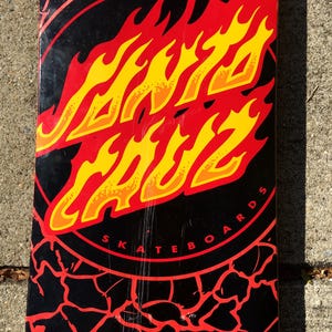 May include: Black skateboard deck with a red and yellow flame design. The deck features the words "SANTA CRUZ" in a flame-like font, surrounded by a red circle with the words "SKATEBOARDS". The background has a cracked pattern.
