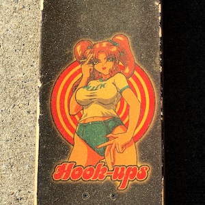 May include: A skateboard with a black grip tape surface featuring a colorful graphic of a woman with red hair, wearing a yellow top and green shorts, with the text "Hook-ups".