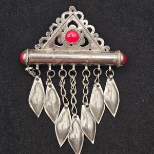May include: A silver-toned triangular brooch featuring an ornate filigree top section with a central red circular stone. A horizontal cylindrical bar with red end caps supports seven dangling silver-toned leaf-shaped charms on metal chains.