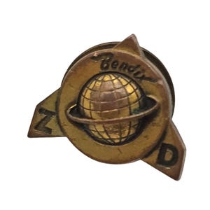 May include: A vintage brass pin with a globe design, encircled by a ring, and the word "Bendix" arched above. The pin has a circular design with stylized "Z" and "D" shapes on either side. The pin is a warm, antique brass colour.