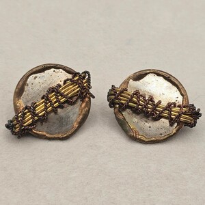May include: A pair of circular earrings featuring a central silver-toned disc with an irregular, textured copper-colored rim. Across the center of each disc, a bundle of thin gold-toned wires is secured by a dark, twisted copper-colored wire wrap.
