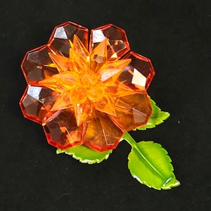 May include: An orange and clear faceted flower brooch with green leaves. The flower has a star-shaped center and a petal design. The leaves are green with yellow highlights. The brooch is set against a black background.