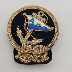 May include: A circular embroidered patch featuring a gold rope border on a black background. The design includes a gold anchor, a nautical knot at the base, and a white and blue flag with green accents. The embroidery uses metallic gold thread.