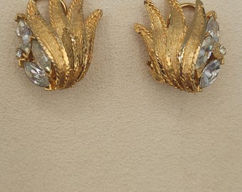 May include: A pair of gold-toned clip-on earrings. Each earring features a textured, stylized leaf design with marquis-cut, light blue gemstones and a small round clear stone. The earrings have a clip-on closure.