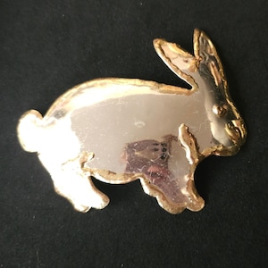 May include: A gold-toned metal bunny pin with a polished silver center. The bunny is in a side profile, with its ears perked up. The edges of the bunny are outlined in gold. The pin is likely vintage and suitable for Easter or spring.