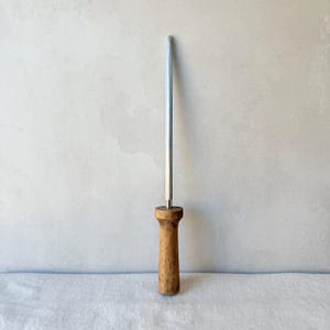 May include: A knife sharpening steel featuring a long, slender, metallic rod with a tapered tip and a cylindrical, light brown wooden handle. The tool is positioned vertically, showcasing the contrast between the smooth metal and the textured wood.