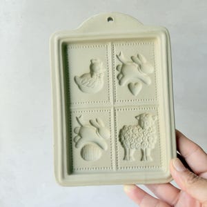 May include: A rectangular, off-white ceramic mold with four Easter-themed designs: a chick, a bunny with a heart, a bunny with an egg, and a sheep. The mold has a small hole at the top for hanging.
