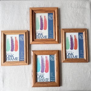 May include: Four rectangular oak frames with a natural wood grain finish. Each frame contains a display insert featuring three vertical brush strokes in mint green, purple, and coral, with the text 5x7 QUALITY OAK FRAME printed in dark blue.