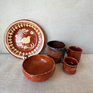 May include: A collection of terracotta pottery including a round plate with a painted bird and geometric border, a large deep bowl, a tall cylindrical cup, and two smaller handled mugs, all featuring earthy red-brown tones and rustic textures.