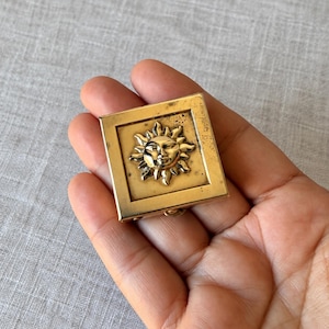 May include: A square gold-toned metal compact case featuring a raised, embossed sun face design in the center. The surface of the metal has a polished finish with visible signs of wear and patina, showing a classic square frame border.