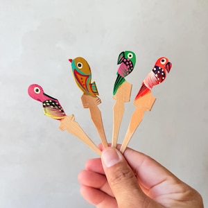 Pode incluir: Uma mão segura quatro palitos de cocktail de madeira em forma de pássaro. Cada palito apresenta um design de pássaro colorido, incluindo vermelho, rosa, amarelo, verde e azul. Os pássaros são pintados com padrões e detalhes intrincados.