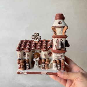 Peut inclure: Une maquette d'église en terre cuite peinte à la main avec un toit en tuiles rouges, un haut clocher et une porte centrale arquée. La structure est ornée de détails peints en blanc, incluant une croix, des motifs géométriques et des figurines.
