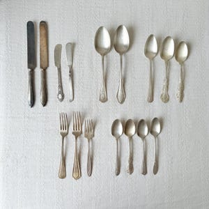 May include: An assortment of antique silverware, including knives, butter knives, forks, and spoons. The silverware is arranged on a white surface, showcasing various designs and patterns. The spoons and forks have different handle styles.