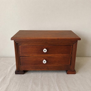 May include: A small rectangular wooden chest featuring two horizontal drawers stacked vertically. Each drawer is equipped with a single round white ceramic knob centred on the front panel. The chest has a dark brown finish and four feet.