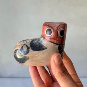 May include: A small, smooth, hand-painted ceramic figurine shaped like a stylized owl. The piece features a reddish-brown head with large, circular eyes and a cream-colored body decorated with black and orange floral and geometric patterns.