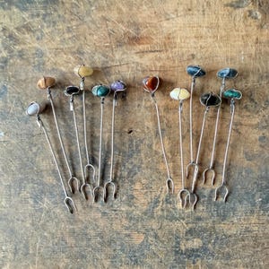 May include: A collection of ten metal appetizer forks, each featuring a unique polished gemstone at the top. The stones vary in color, including shades of brown, green, purple, black, and white, all secured with silver-toned wire.