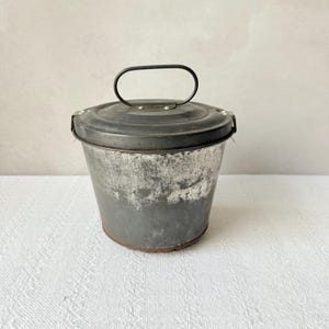 Könnte beinhalten: Ein kleiner, alter Metallbehälter mit Deckel und Griff. Der Eimer ist grau mit Gebrauchsspuren, was ihm ein rustikales Aussehen verleiht. Der Deckel ist dunkelgrau und hat einen einfachen, gebogenen Griff. Ein charmantes Dekorationsstück.