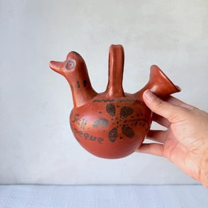 May include: A terracotta-coloured ceramic vessel with a bird-like design. The vessel features a spout, a handle, and a bird's head with an eye. The body is decorated with dark brown floral patterns and text. A unique piece of pottery.