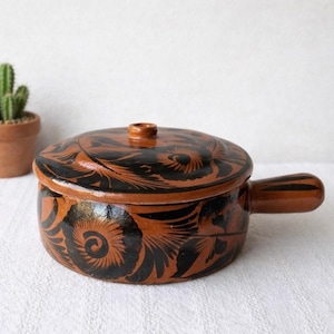 Op de afbeelding: Een ronde, terracotta-kleurige keramische pot met een bijpassend deksel en een enkele handgreep aan de zijkant. Het buitenoppervlak is voorzien van een handgeschilderd zwart bloemmotief met wervelende details op het deksel en de pot.
