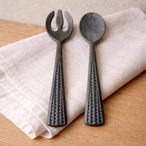 May include: A dark gray salad fork and spoon set. The fork has three tines, and the spoon has a rounded bowl. Both utensils have textured handles and are resting on a beige linen napkin, placed on a wooden surface.