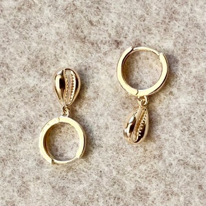 Peut inclure: Boucles d'oreilles créoles dorées avec un charm en forme de coquillage pendant. Les boucles d'oreilles ont une finition polie et un design simple et élégant. Les breloques coquillages sont détaillées.