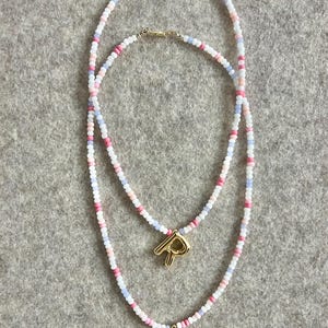 Könnte beinhalten: Zwei übereinanderliegende Halsketten mit rosa, weißen und blauen Perlen. Die Halsketten haben goldene Buchstabenanhängern, einer ist ein 'R' und der andere ist ein 'L'.