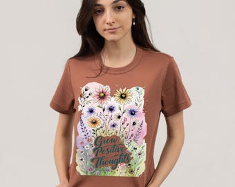 May include: A brown t-shirt with a floral design and the text "Grow Positive Thoughts." The design features colourful flowers and foliage. The shirt is made of a soft material.