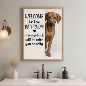 Puede incluir: Un cuadro enmarcado con el texto "WELCOME to the BATHROOM" y "a Ridgeback will be with you shortly." Un perro Rhodesian Ridgeback marrón es parcialmente visible. El marco es de color marrón claro.