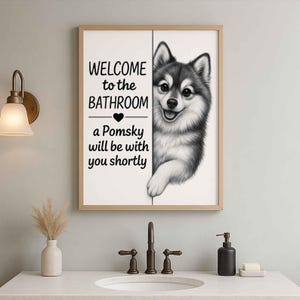 Pode incluir: Uma impressão emoldurada com o texto "WELCOME to the BATHROOM" e "a Pomsky will be with you shortly." A impressão apresenta uma ilustração a preto e branco de um cão Pomsky espreitando por trás de uma parede. A moldura é castanha clara.