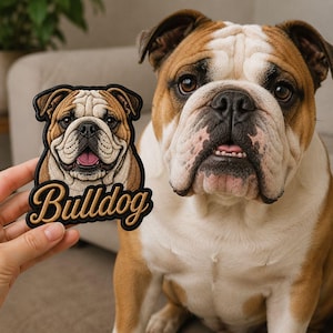 May include: A fabric patch featuring an embroidered illustration of a brown and white bulldog face with the word Bulldog written in cursive script below the image, displayed next to a live brown and white bulldog with similar facial markings.