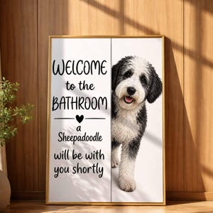 May include: A framed print with a gold border features a black and white Sheepadoodle dog. The text reads "WELCOME to the BATHROOM a Sheepadoodle will be with you shortly". The dog is smiling with its tongue out.