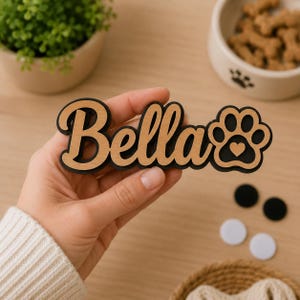 Pu&ograve; includere: Una targhetta in legno personalizzata con il nome Bella in carattere corsivo, sovrapposta a uno sfondo nero. Il design include l'icona di un'impronta di zampa con un piccolo cuore ritagliato, realizzata in legno chiaro e materiale nero.