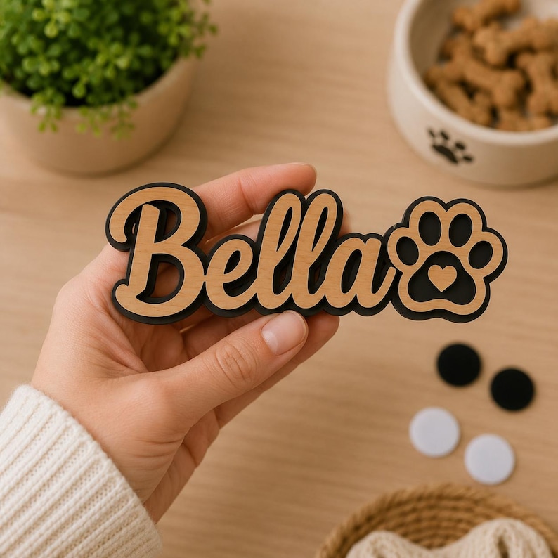 Pu&ograve; includere: Una targhetta in legno personalizzata con il nome Bella in carattere corsivo, sovrapposta a uno sfondo nero. Il design include l'icona di un'impronta di zampa con un piccolo cuore ritagliato, realizzata in legno chiaro e materiale nero.