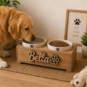 Pu&ograve; includere: Un supporto rettangolare in legno per animali domestici con due ciotole in ceramica bianca. La parte anteriore del supporto presenta il nome Bella in lettere corsive di legno scuro accanto a un'icona a forma di zampa nera.