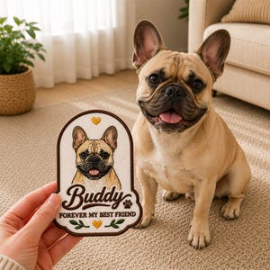 May include: A tan French Bulldog sits next to a patch held by a hand. The patch features a portrait of a French Bulldog with the name "Buddy" and the text "Forever My Best Friend". The patch has a brown border and decorative elements.