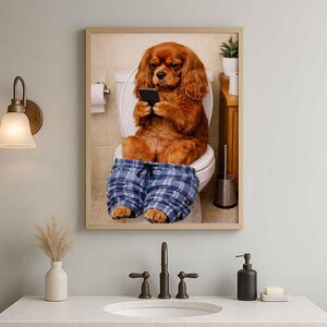 May include: A framed print depicts a Cavalier King Charles Spaniel sitting on a toilet, engrossed in a smartphone. The dog wears blue plaid pajama pants. The bathroom setting includes a toilet paper roll and a small plant.