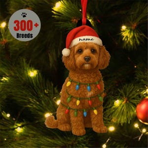 May include: A brown dog Christmas ornament wearing a Santa hat with the word "name" and a string of colorful lights. The ornament is hanging in front of a Christmas tree. A circular graphic reads "300+ Breeds".