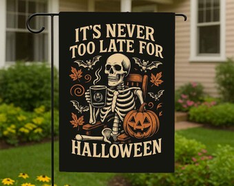 May include: A black rectangular garden flag featuring a skeleton sitting in a chair, holding a coffee mug, and a jack-o'-lantern. The text reads IT'S NEVER TOO LATE FOR HALLOWEEN. The design includes orange autumn leaves and small bats.