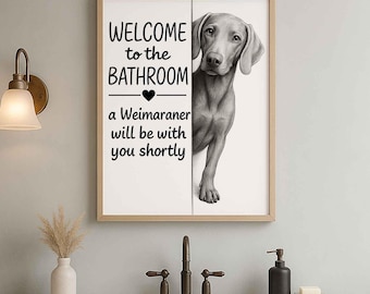 May include: Framed bathroom sign with a Weimaraner dog illustration. The sign reads "WELCOME to the BATHROOM" above a heart symbol, followed by "a Weimaraner will be with you shortly" in black text. The artwork is in a light-colored wooden frame.