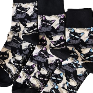 May include: A set of three crew socks featuring a repeating pattern of black cats with large eyes on a cream background. Each sock has a solid black cuff, heel, and toe section. The word "harmonia" is printed in small white text on each sock.