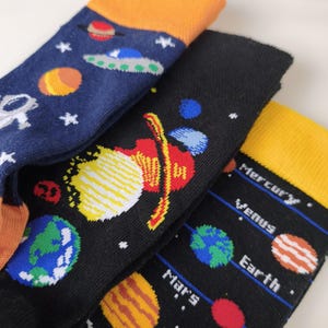 May include: Three pairs of crew socks featuring space-themed designs. One pair is navy blue with orange cuffs, planets, and a UFO. The second is black with colourful planets. The third is black with yellow cuffs, labelled planets, and stars.