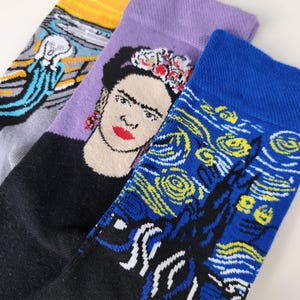 May include: Three pairs of crew socks featuring famous art prints. One sock displays The Scream with a yellow sky, another features a portrait of Frida Kahlo with a floral headpiece on a lavender background, and the third shows Starry Night.
