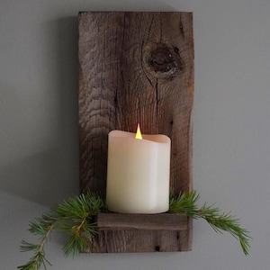 May include: A rustic wooden wall sconce with a cream-colored candle and a faux flickering flame. The candle sits on a small shelf, and is decorated with green pine branches. The wooden plank has a natural, weathered appearance.