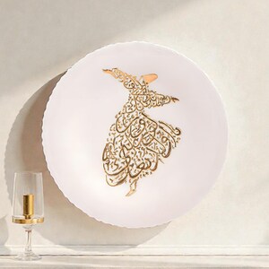 Puede incluir: Un plato decorativo blanco con borde festoneado, con un diseño de caligrafía dorada de una figura giratoria. También se ven un candelabro con detalles dorados y una ramita de flores amarillas.