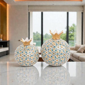 Puede incluir: Dos esculturas decorativas de cerámica con forma de granada. Los cuerpos esféricos están adornados con un patrón repetido de motivos florales azules y amarillos sobre un fondo blanco. Cada una tiene una parte superior dorada en forma de corona. Las esculturas se exhiben sobre una superficie de mármol.