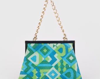 Puede incluir: Un bolso de mano de estilo vintage con un patrón geométrico en tonos azules y verdes. El bolso tiene un asa de cadena dorada y un marco negro. El diseño presenta formas de diamantes y cuadrados.